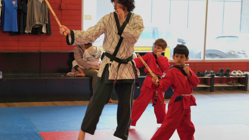 A martial arts instructor in a white uniform teaching two young students in red uniforms how to use traditional wooden sticks at Yuen's Family Martial Arts in Coquitlam