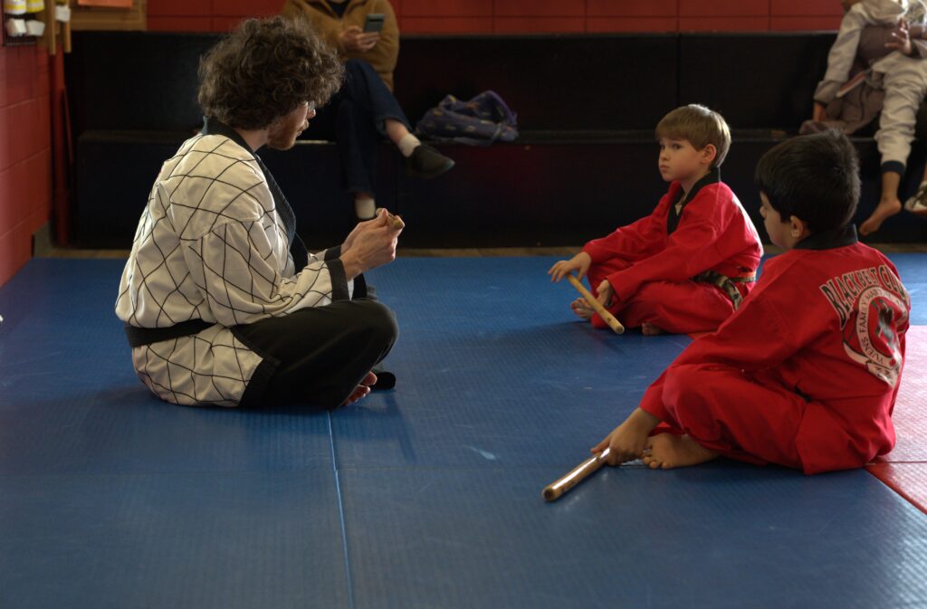 A group of young students sitting respectfully and listening as an instructor explains the fundamentals of Filipino Martial Arts (FMA) stick training.