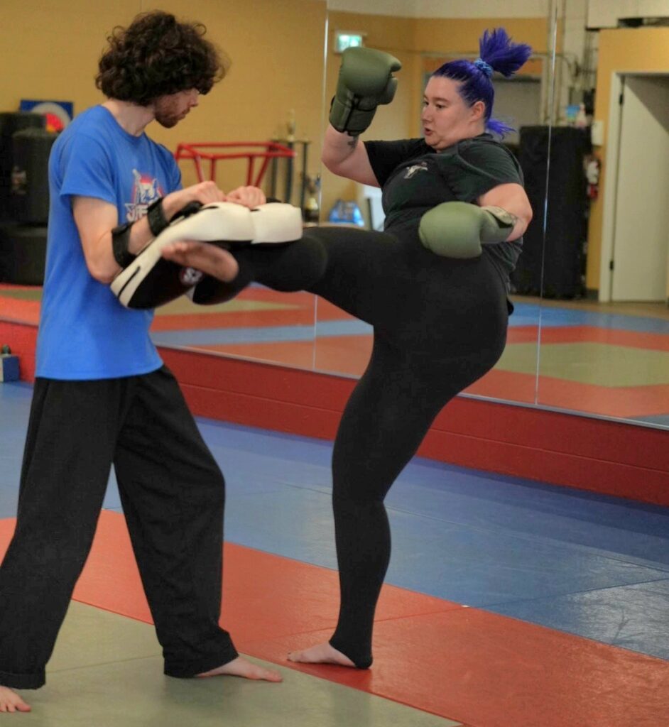 A female student practicing a high power kickboxing technique on target pads with a coach during a beginner-friendly adult martial arts class in Coquitlam.