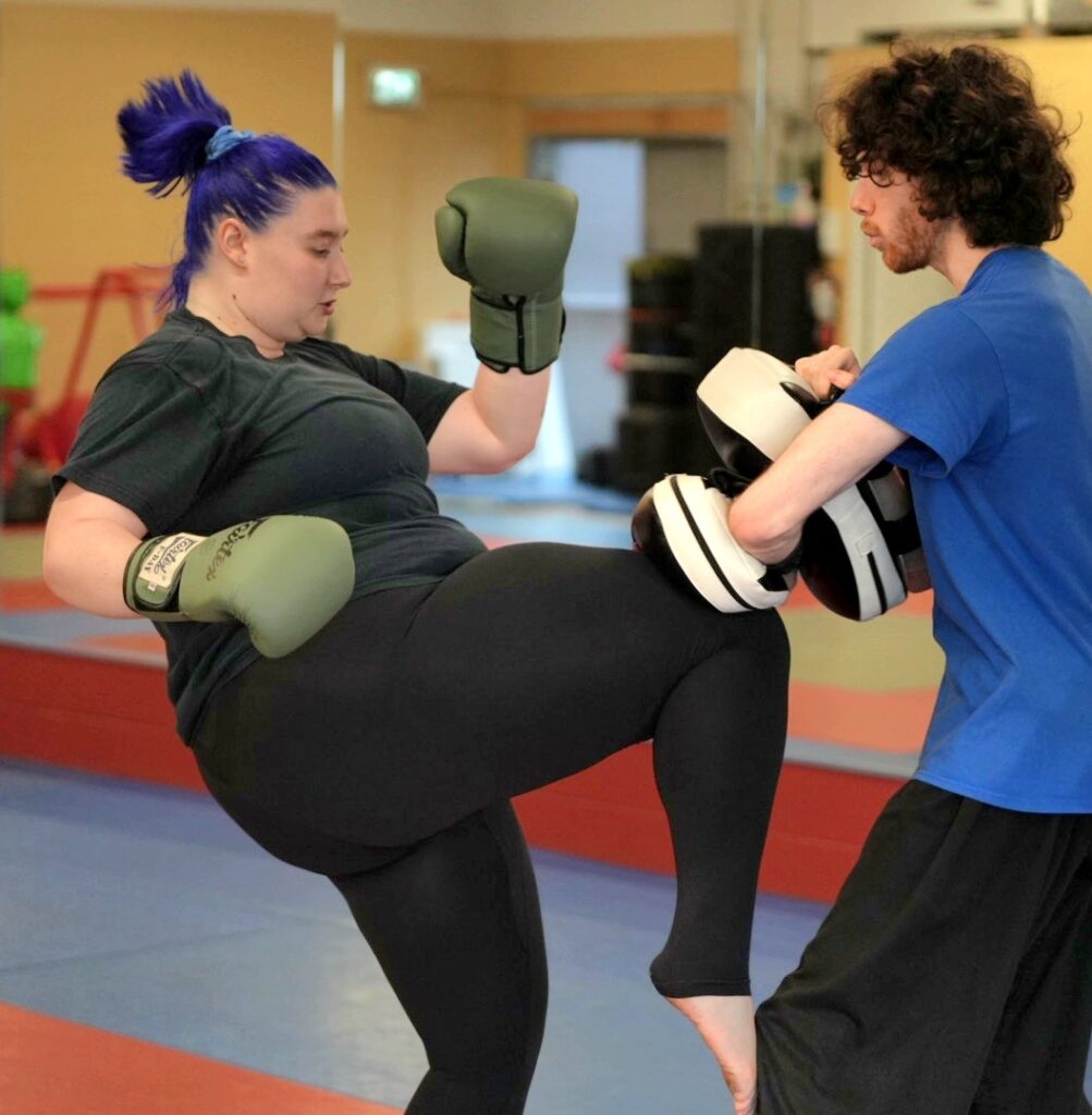 An adult student practicing an introductory Muay Thai knee strike on curved pads held by a supportive coach at Yuen's Family Martial Arts in Coquitlam.